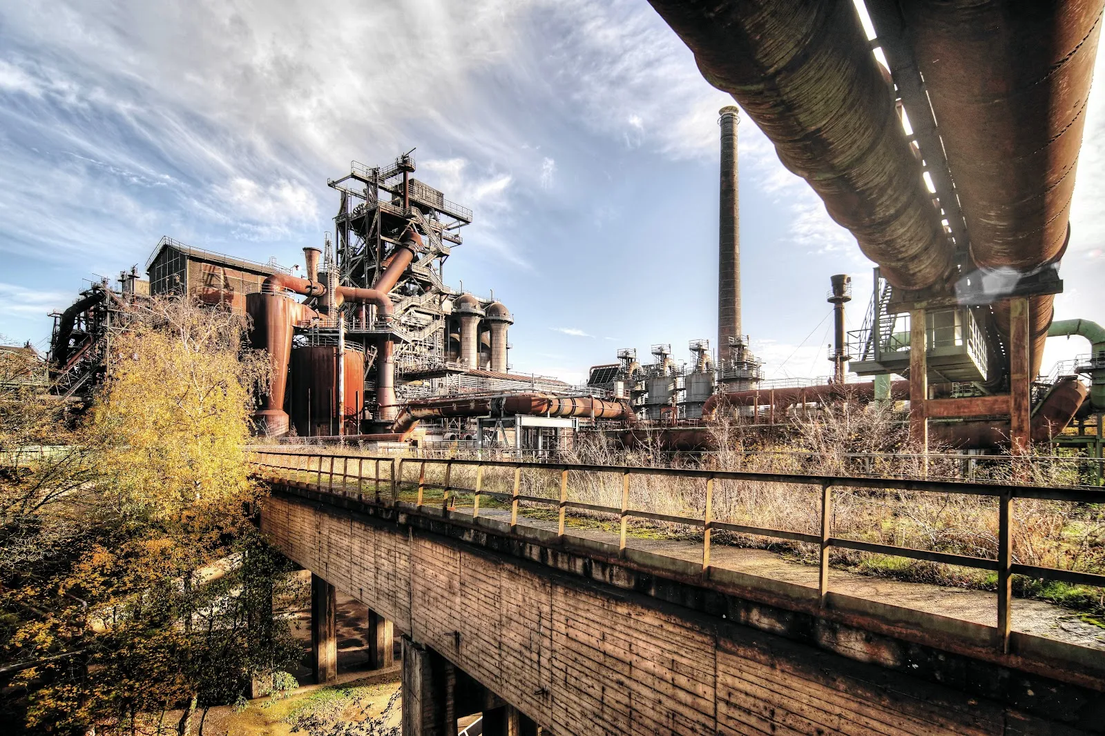 Люди гуляют по Ландшафтному парку Дуйсбург-Норд среди старых промышленных сооружений и зелени.