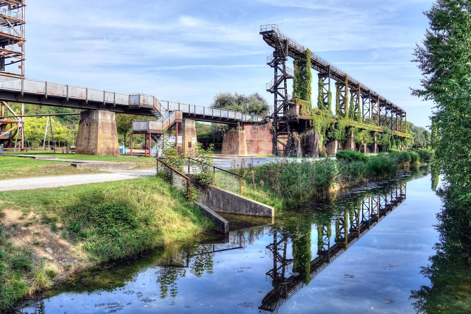 Люди гуляют по Ландшафтному парку Дуйсбург-Норд среди старых промышленных сооружений и зелени.