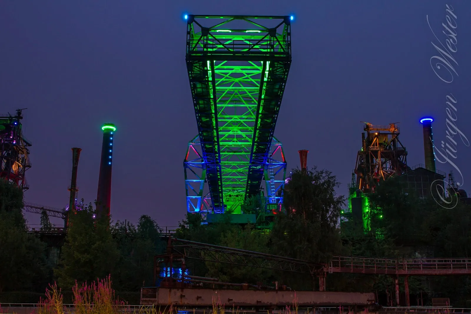 Люди гуляют по Ландшафтному парку Дуйсбург-Норд среди старых промышленных сооружений и зелени.