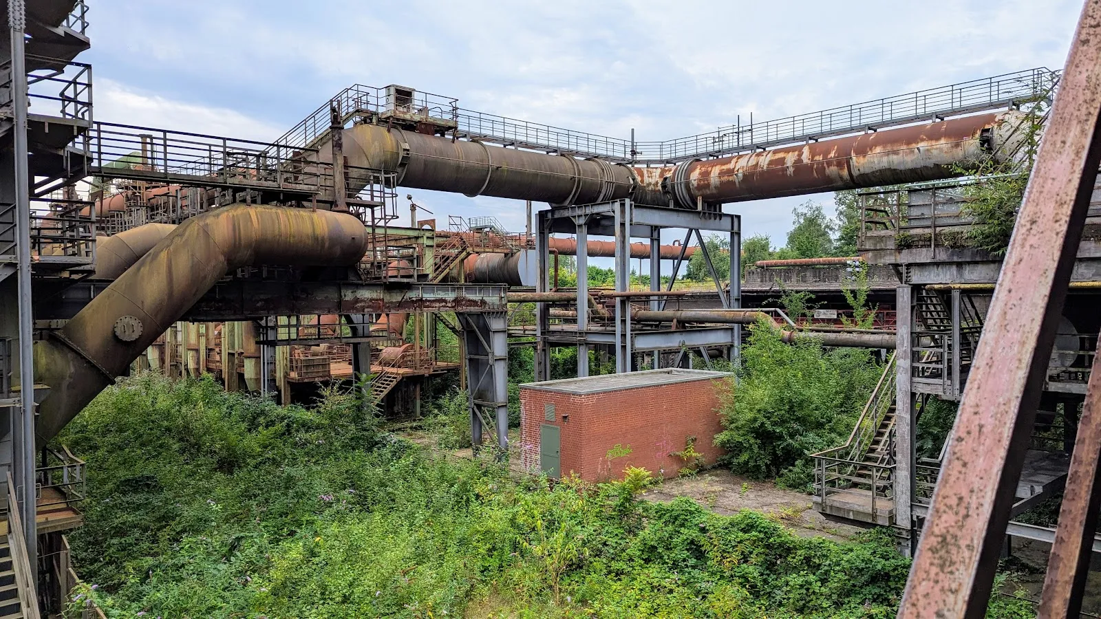 Люди гуляют по Ландшафтному парку Дуйсбург-Норд среди старых промышленных сооружений и зелени.
