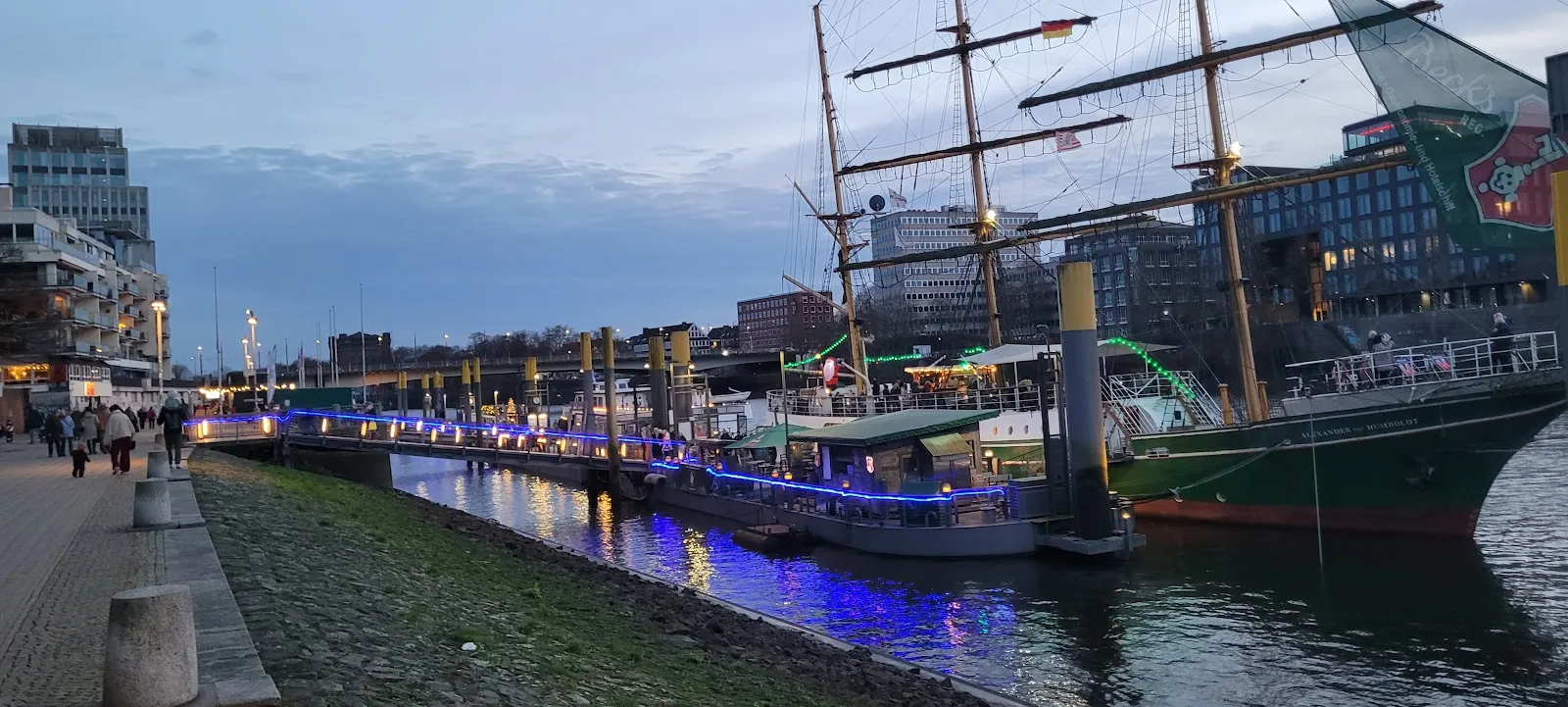 Люди гуляют по набережной Шлахте в Бремене, Германия, с магазинами и кафе.